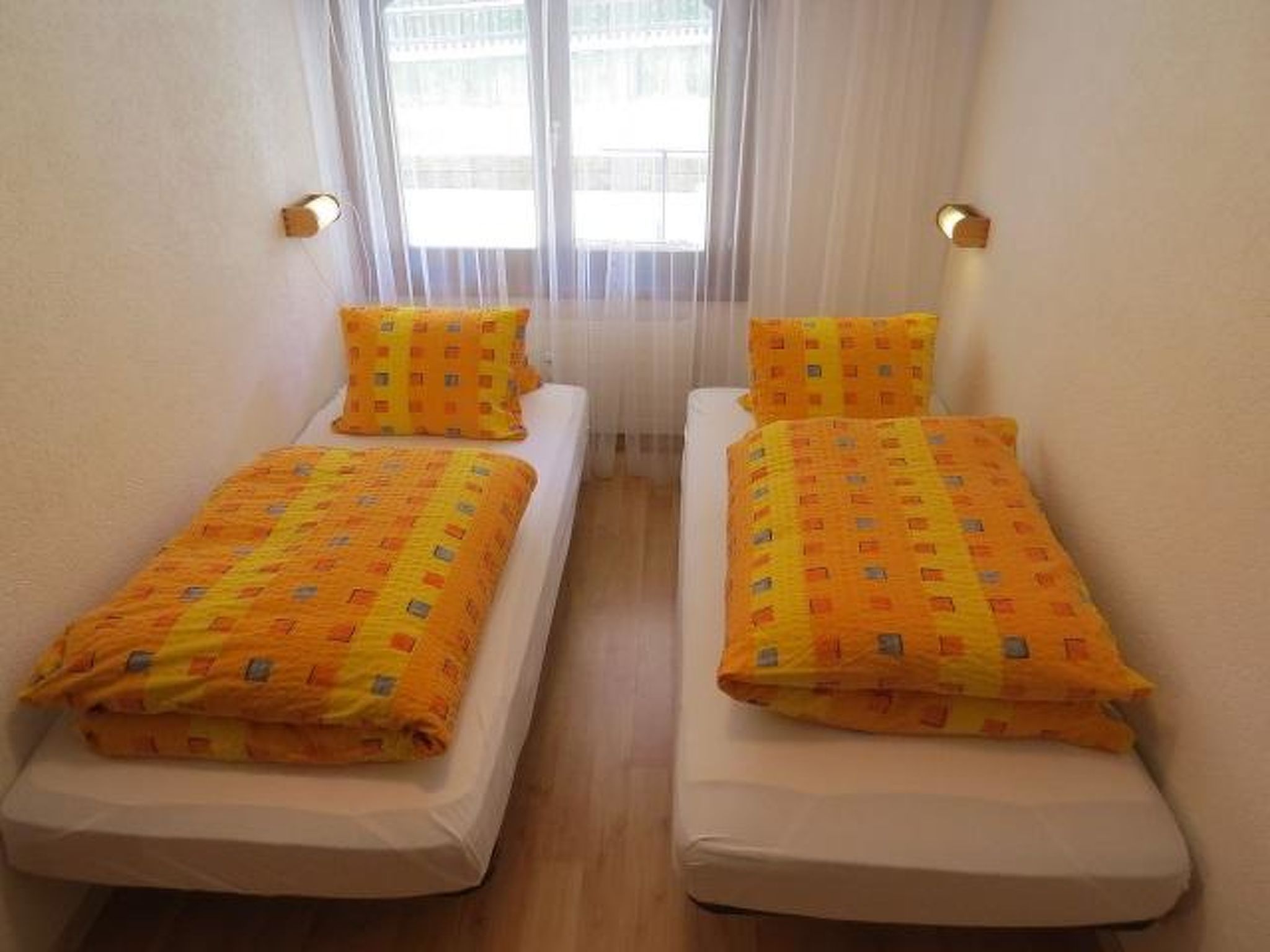 A room with two single beds, each with a patterned blanket and pillow, a window with sheer curtains, and a wall-mounted light fixture.