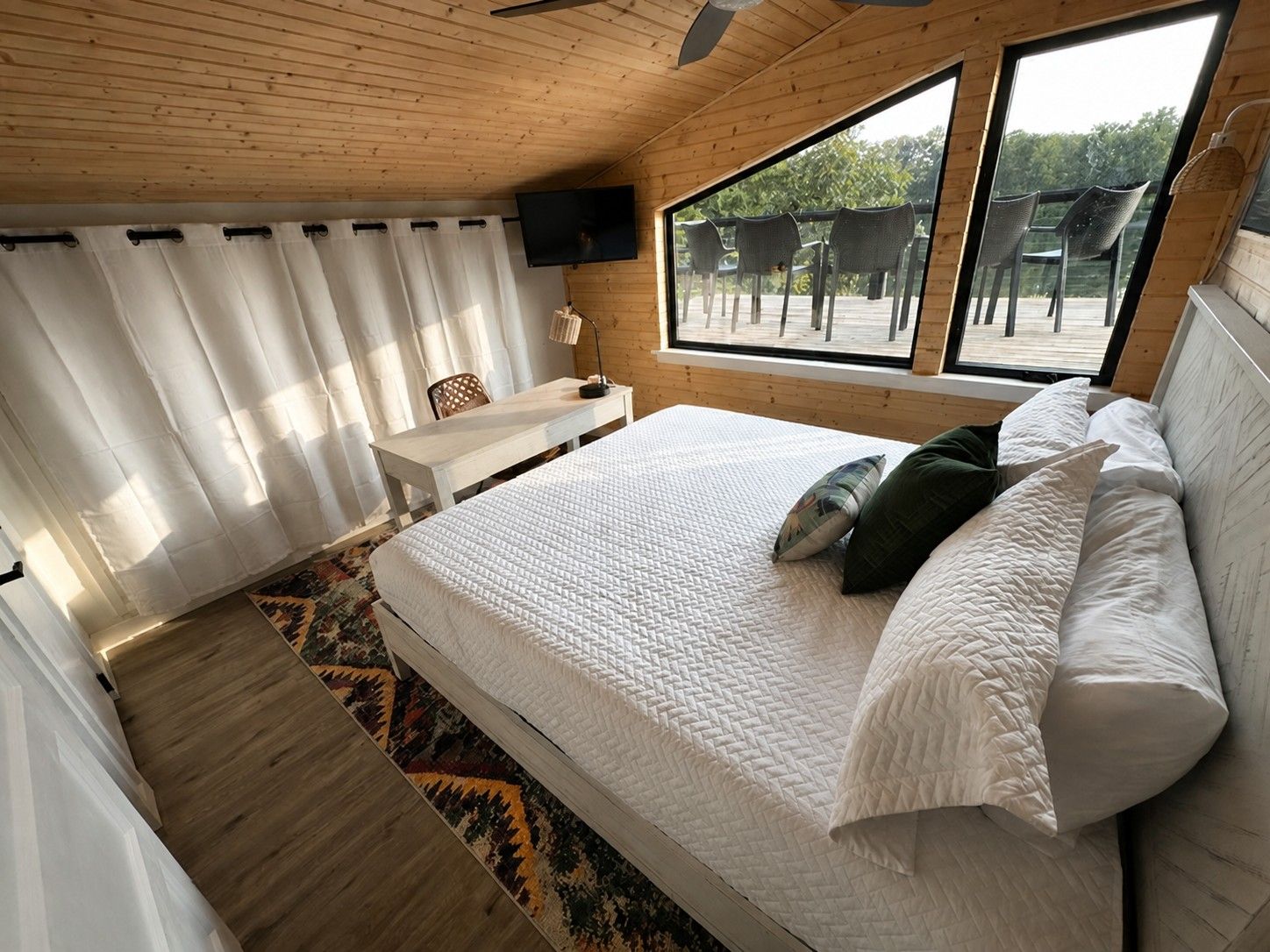 Ein Schlafzimmer mit einem großen Bett, einem Schreibtisch, einem Stuhl und einem Fernseher. Es gibt Fenster mit Blick ins Freie.