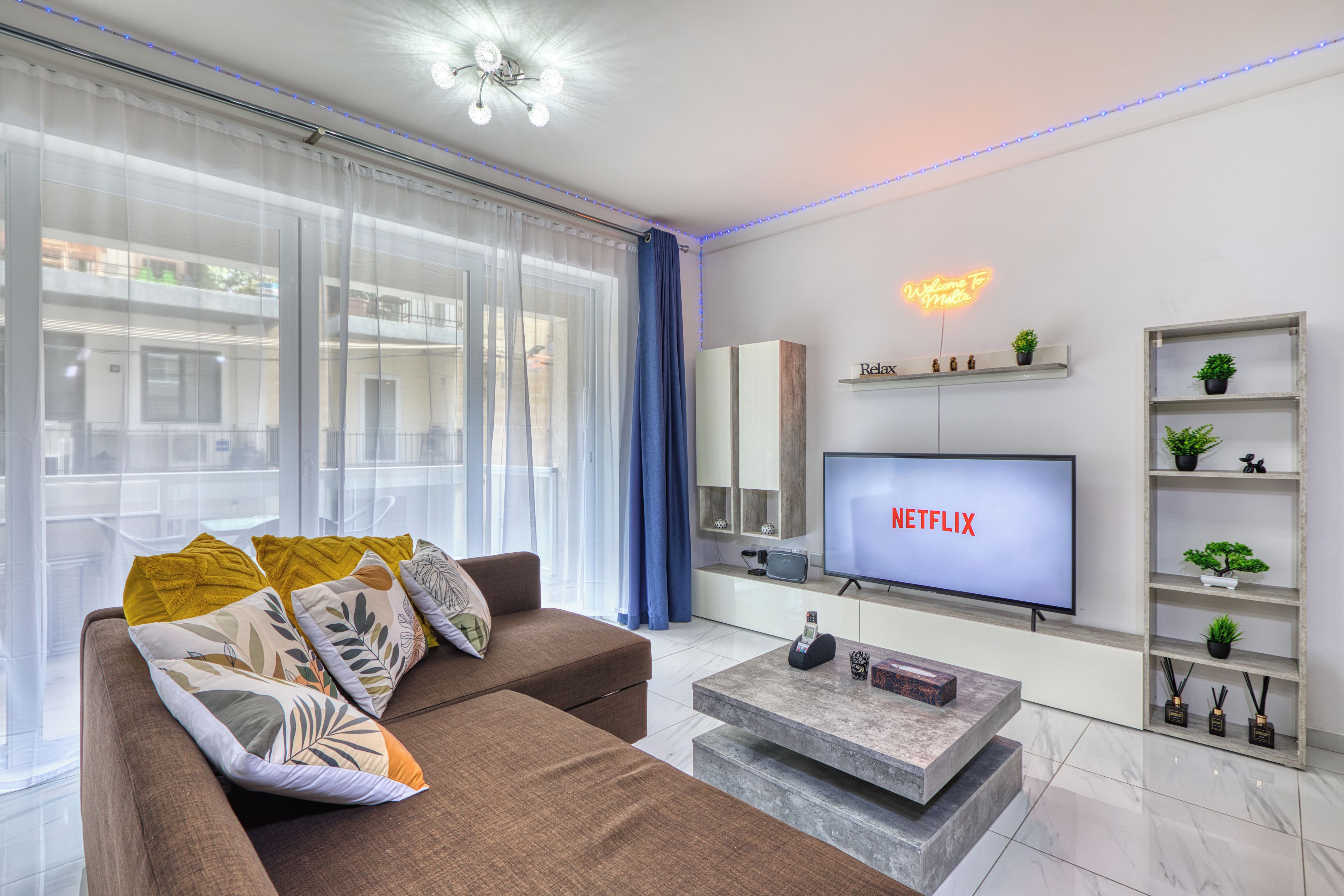 A modern living room with a flat-screen TV, a sofa, a coffee table, and a decorative shelf with plants.