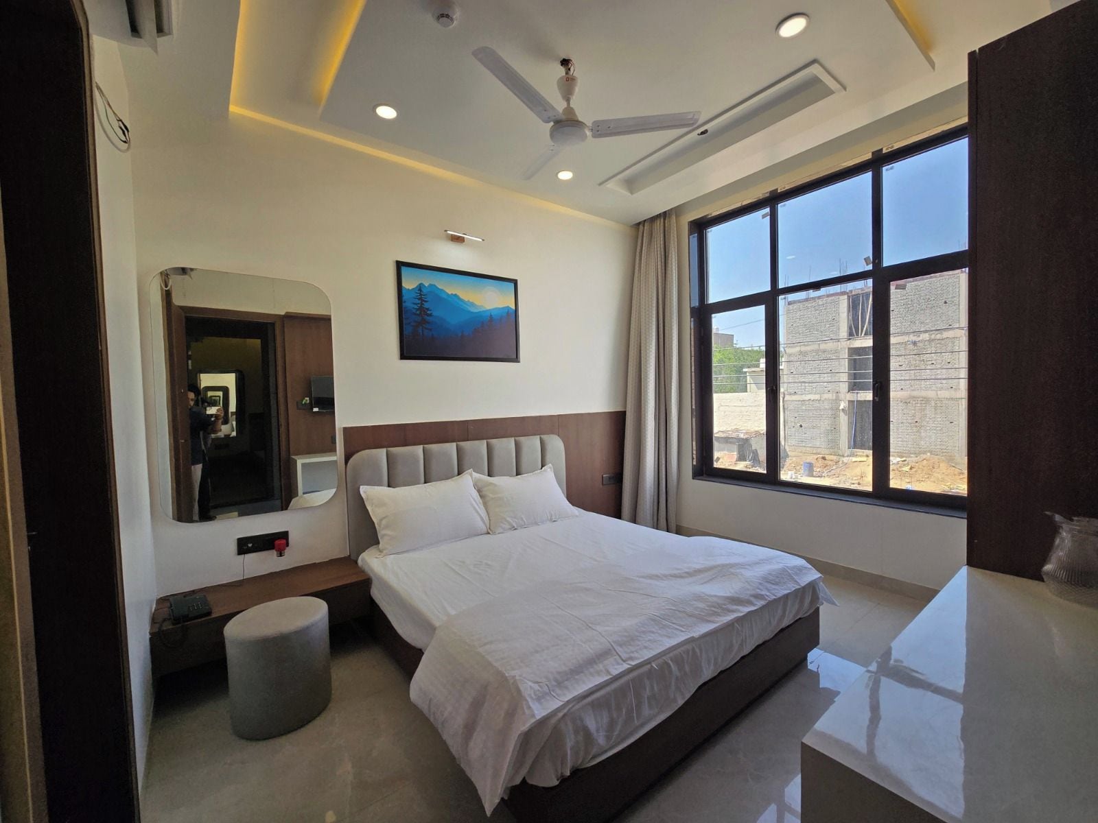 A hotel room with a bed, a bedside table, a mirror, a window with a view of the outside, and a ceiling fan.