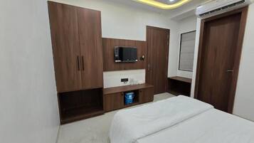 A bedroom with a bed, a TV mounted on a wooden cabinet, a small shelf, and a door with a window.