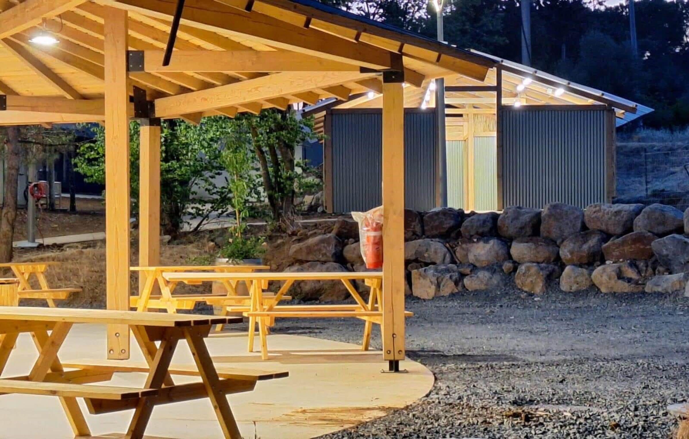 A wooden pavilion with picnic tables, a stone wall, and a metal fence in the background.