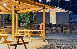 Un pabellón de madera con mesas de picnic, un muro de piedra y una valla metálica al fondo.