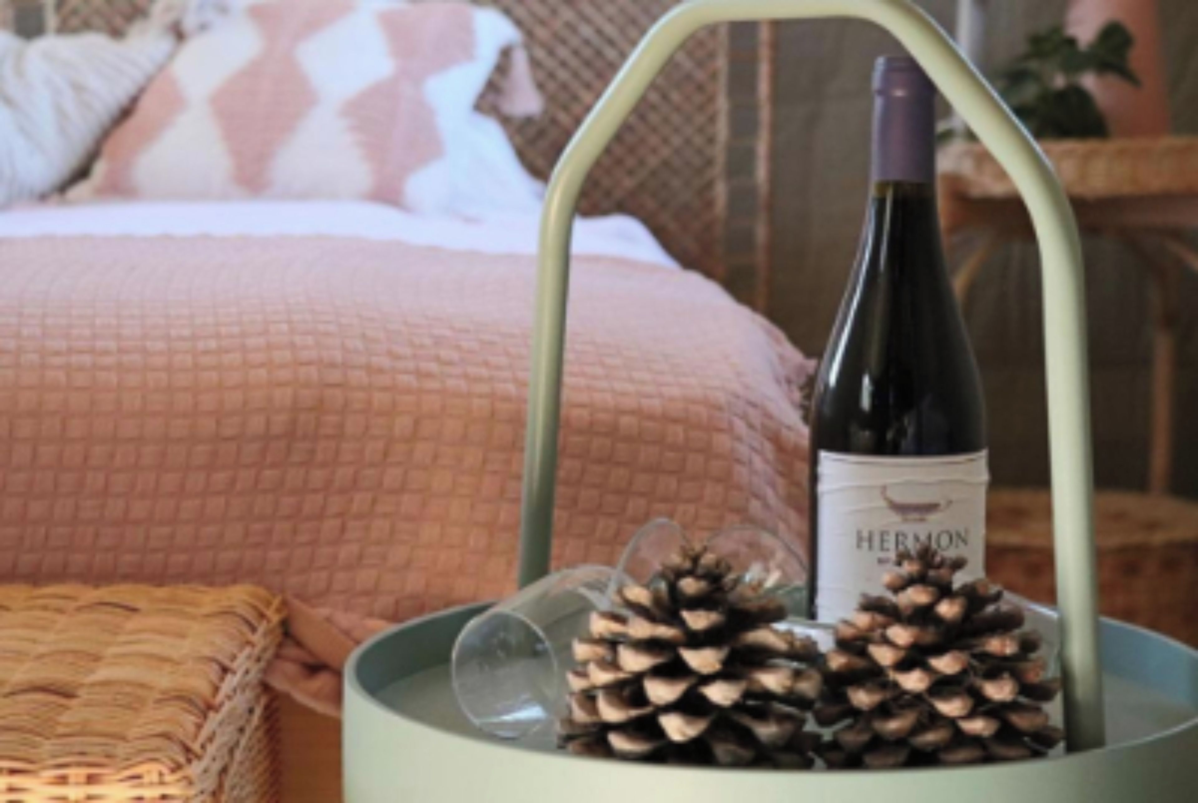 Una cama con colcha rosa, una cesta de mimbre, una botella de vino y piñas.