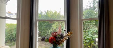 A table with a vase of flowers in front of a window with a view of greenery outside.