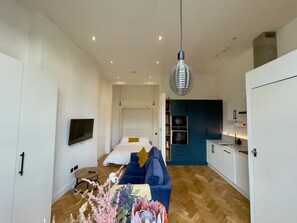 Ein modernes Wohnzimmer mit einer Couch, einem Fernseher und einem Essbereich, im Hintergrund ist eine Küche zu sehen.
