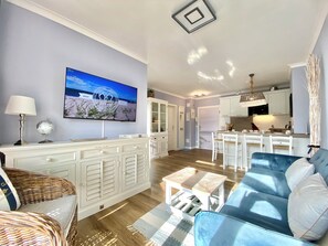 A modern living room with a large flat-screen TV, a dining area with white chairs, and a kitchen with white cabinets. - Ostseestrand-23, prime beachfront location, balcony, elevator, sauna (Kühlungsborn)