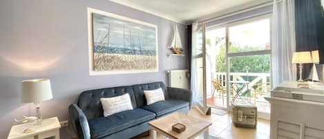 Una sala de estar con un sofá azul, una mesita auxiliar blanca, una mesa de centro, un cuadro con una escena de playa y vistas al exterior a través de un ventanal.