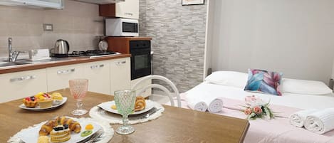 A kitchen with a microwave, stove, and sink, a dining table with pastries and a glass, and a bed with a pillow and rolled towels.