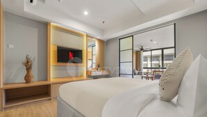 A modern hotel room with a large bed, a TV mounted on a wooden panel, a small table, and a view of the outside through large windows. - Naya by Kozystay - Nusa Dua (Kuta)