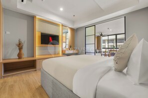 A modern hotel room with a large bed, a TV mounted on a wooden panel, a small table, and a view of the outside through large windows. - Naya by Kozystay - Nusa Dua (Kuta)