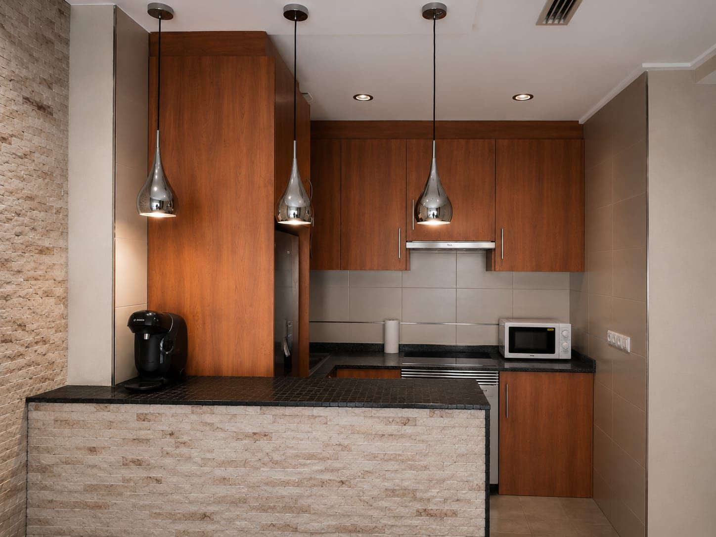 A modern kitchen with wooden cabinets, a built-in island, pendant lights, and a microwave.