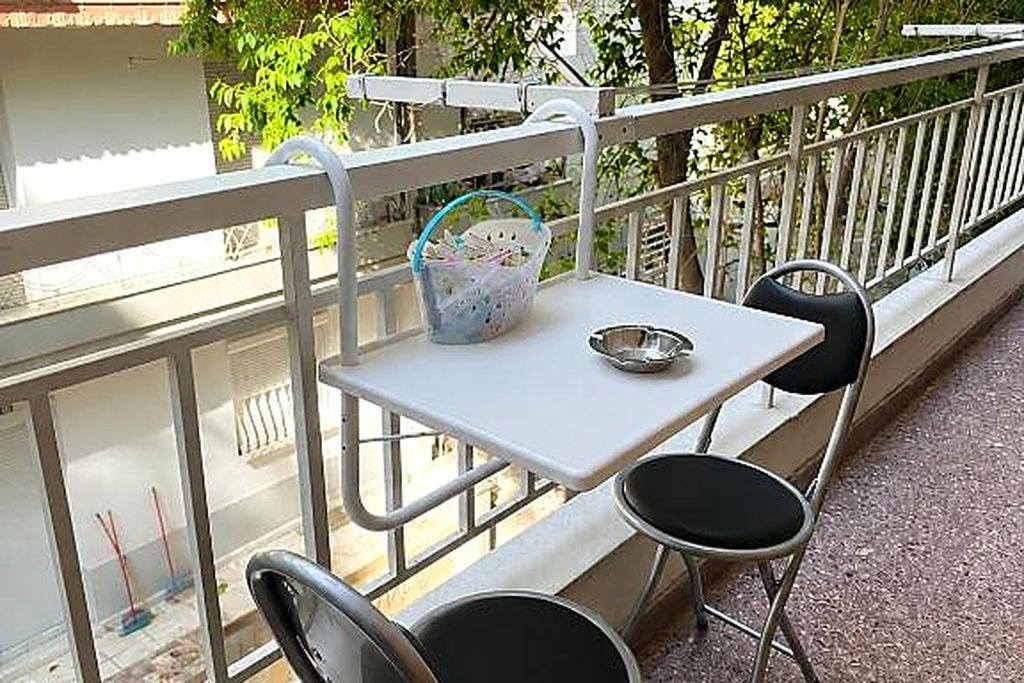 Un balcon avec une table blanche et deux chaises noires, un panier contenant divers objets et une rambarde en métal.