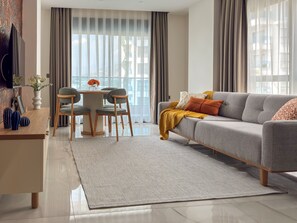 A modern living room with a grey sofa, a dining table with chairs, and a large window with curtains. - Qoople Green Ocean C49 - Cozy 1-bedroom apartment in Alanya (Alanya)