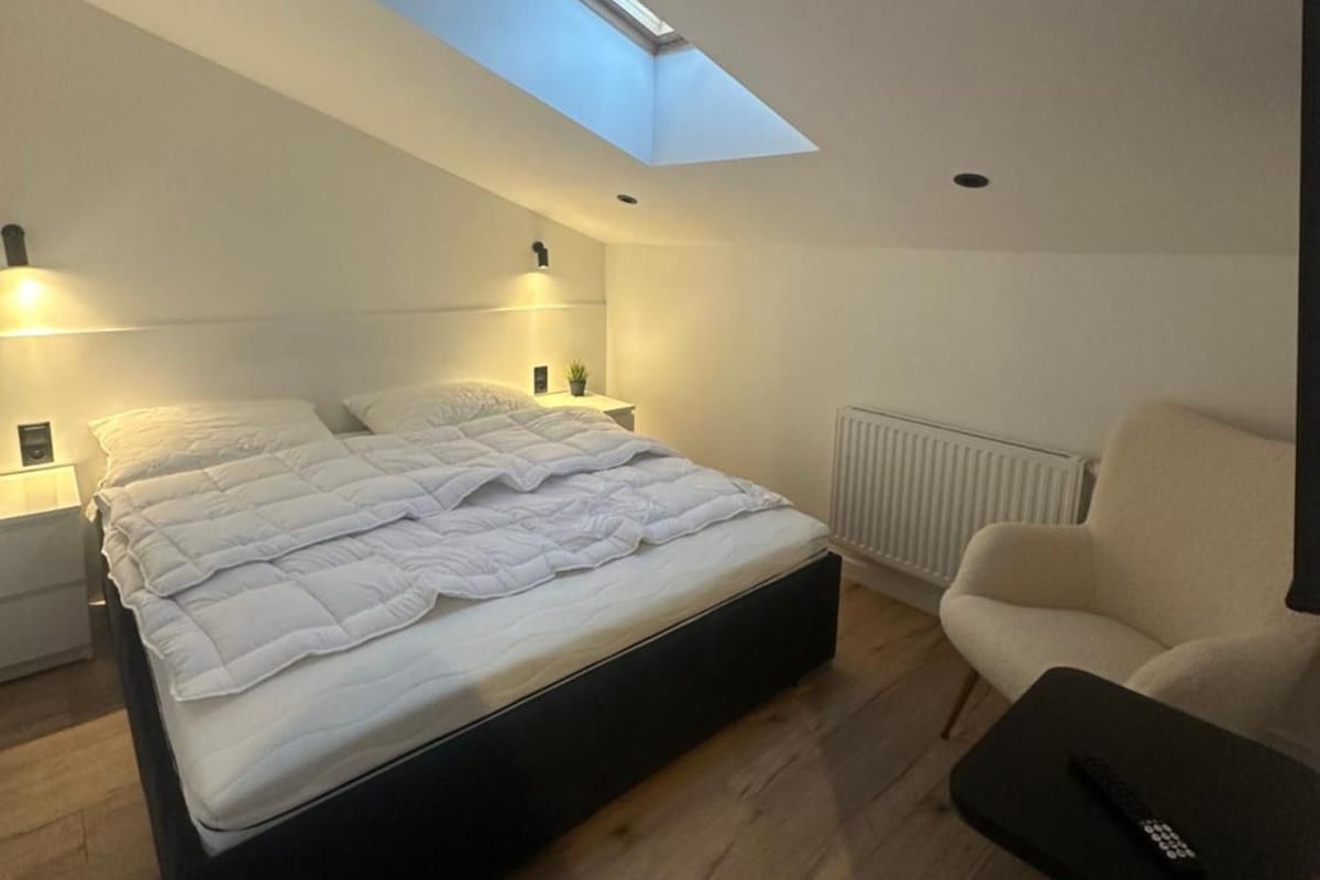 Un dormitorio moderno con tragaluces, una cama, lámparas de noche, un radiador y una silla.