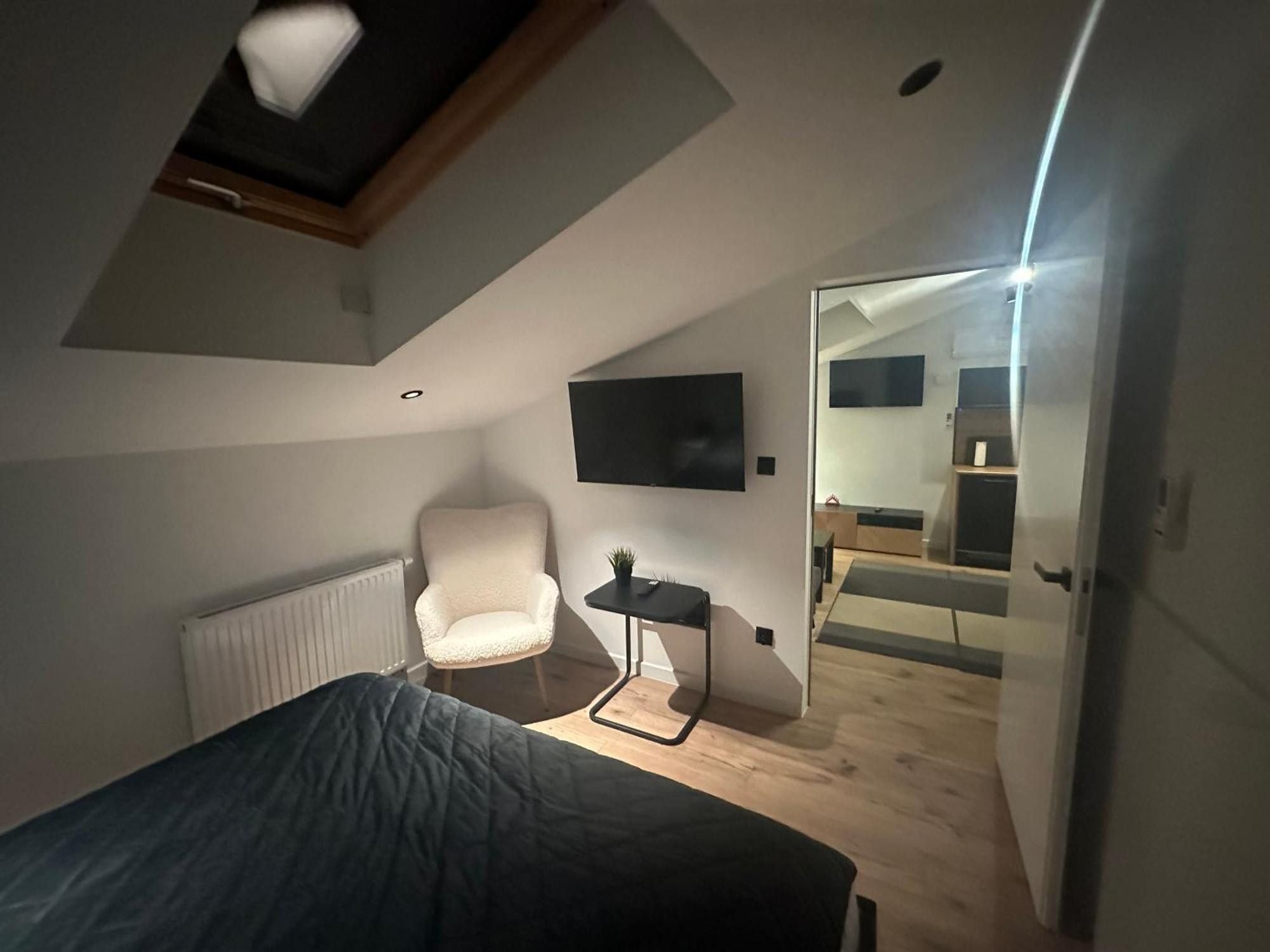 A bedroom with a sloped ceiling, a bed, a chair, a small table with a plant, and a TV mounted on the wall.