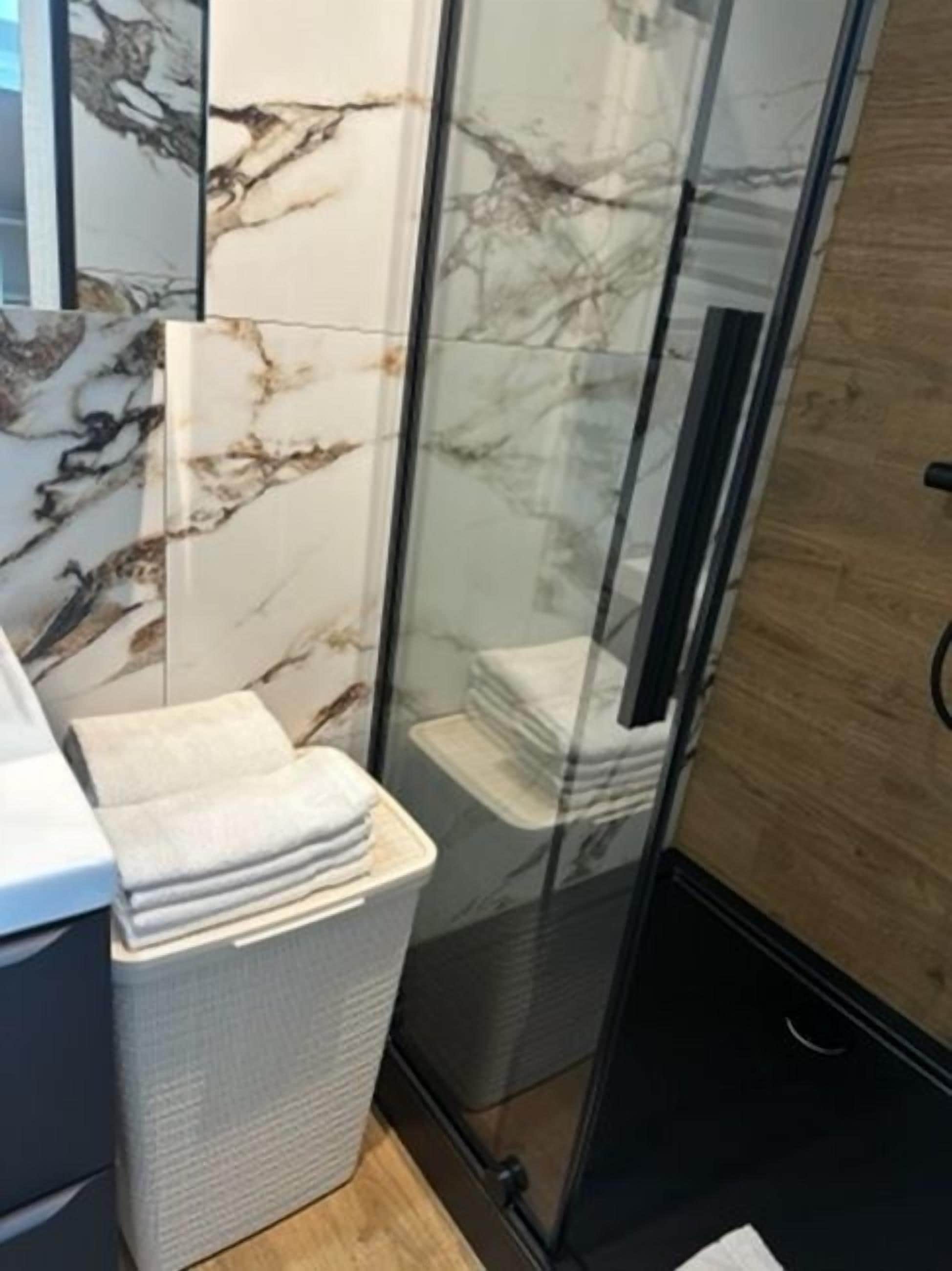 A bathroom with a glass shower enclosure, a marble wall, and a towel rack with folded towels.