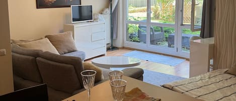 A modern living room with a sofa, a dining table with wine glasses, a TV, and a large window with a view of greenery.