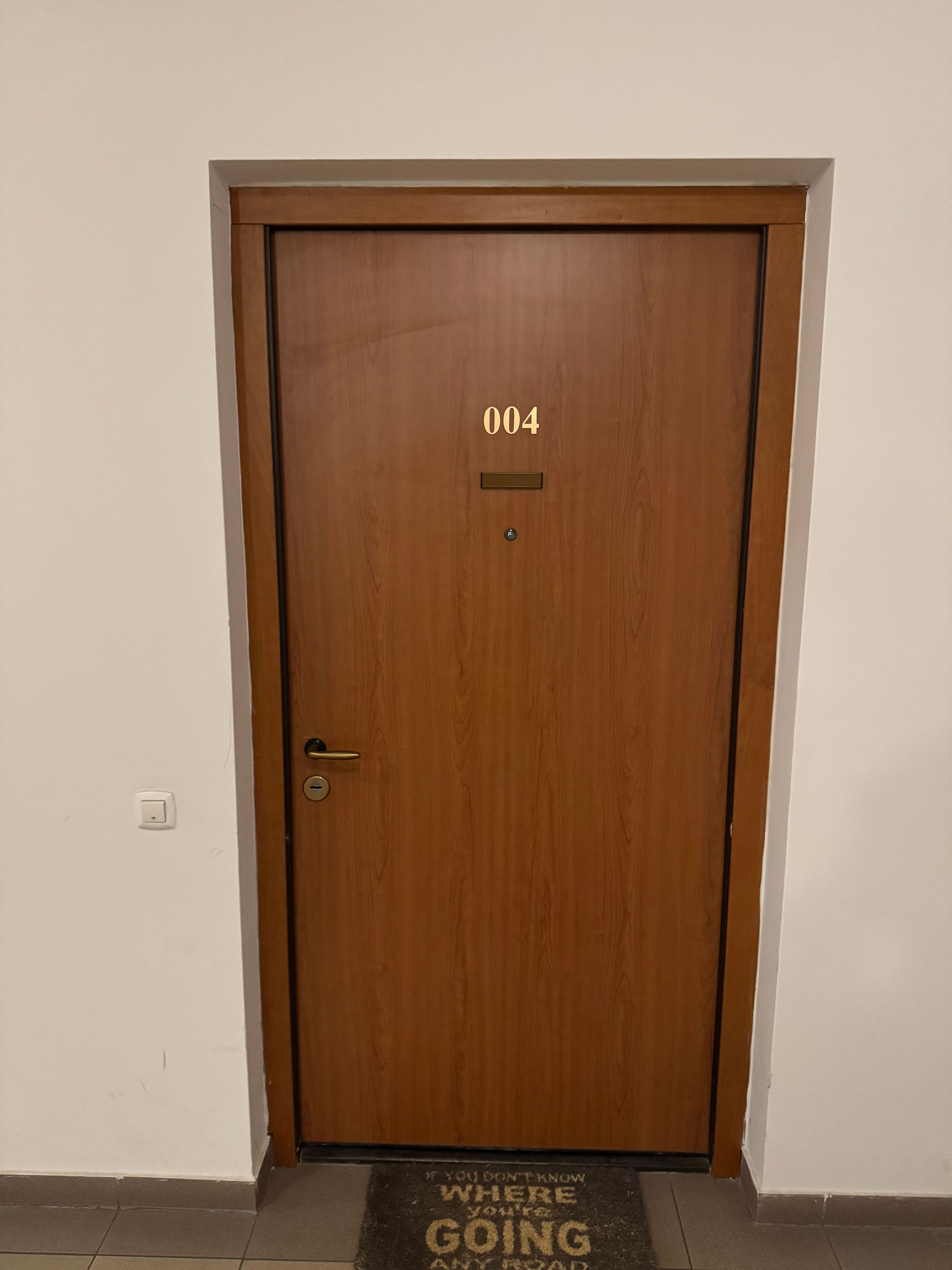Egy fa ajtó, rajta a 004-es szám, egy csengő és egy üdvözlő felirattal ellátott lábtörlő.