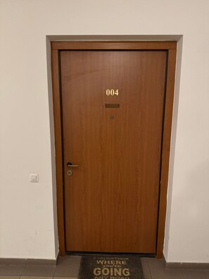 Eine Holztür mit der Nummer 004, einem Klingelknopf und einer Fußmatte mit Aufschrift.