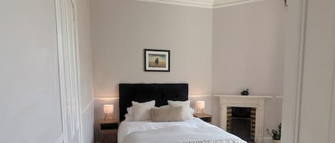 Un dormitorio con cama, mesitas de noche, chimenea y un cuadro en la pared.