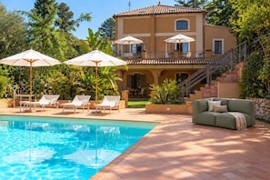 A swimming pool with lounge chairs and umbrellas, a green sofa, and a two-story building with a balcony. - Capovillas - Silent Country House (Gasponi)
