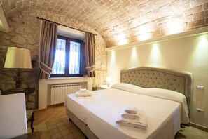 A hotel room with a stone wall, a large bed with a headboard, a window with curtains, a radiator, and a table with a lamp. - Capovillas - Silent Country House (Gasponi)