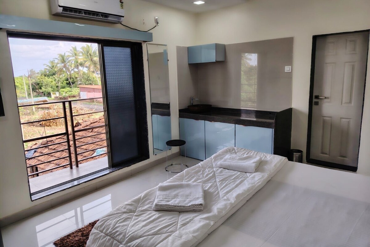 A hotel room with a bed, a small kitchenette, a bathroom, and a balcony with a view of the outdoors.