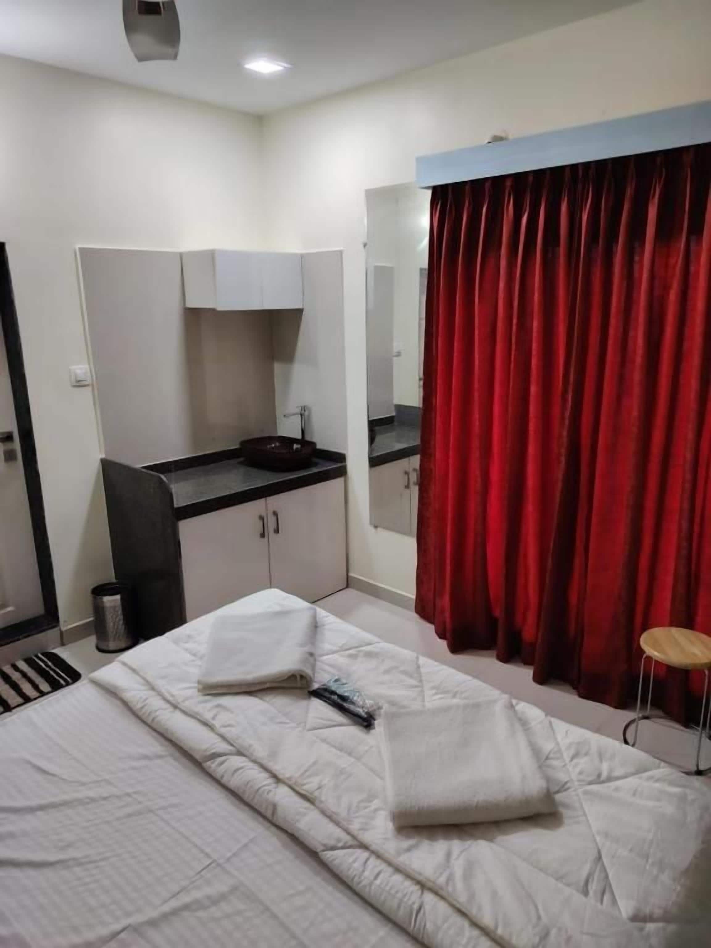 A hotel room with a bed, a desk with a sink, a mirror, and a red curtain.