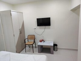 A bedroom with a bed, a wardrobe, a desk with a chair, a television mounted on the wall, and a trash bin.