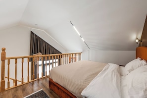 Un dormitorio con techo inclinado, una escalera de madera, una cama con ropa de cama blanca y una mesita de noche.