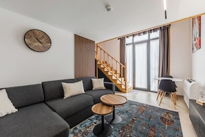 Una sala de estar moderna con un sofá gris, una mesa de centro redonda de madera y una escalera que conduce a un piso superior.