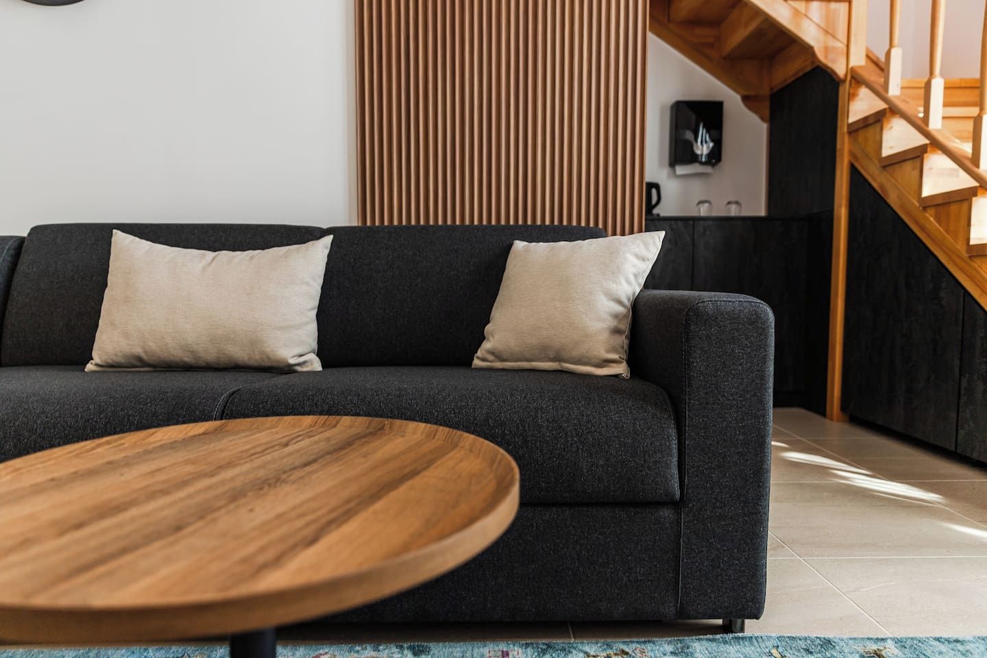 En moderne stue med en mørkegrå sofa, to beige puder og et rundt træbord.