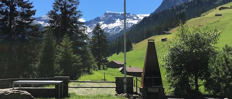 Un asta con una bandera, una mesa de picnic, una parrilla y un paisaje montañoso al fondo.
