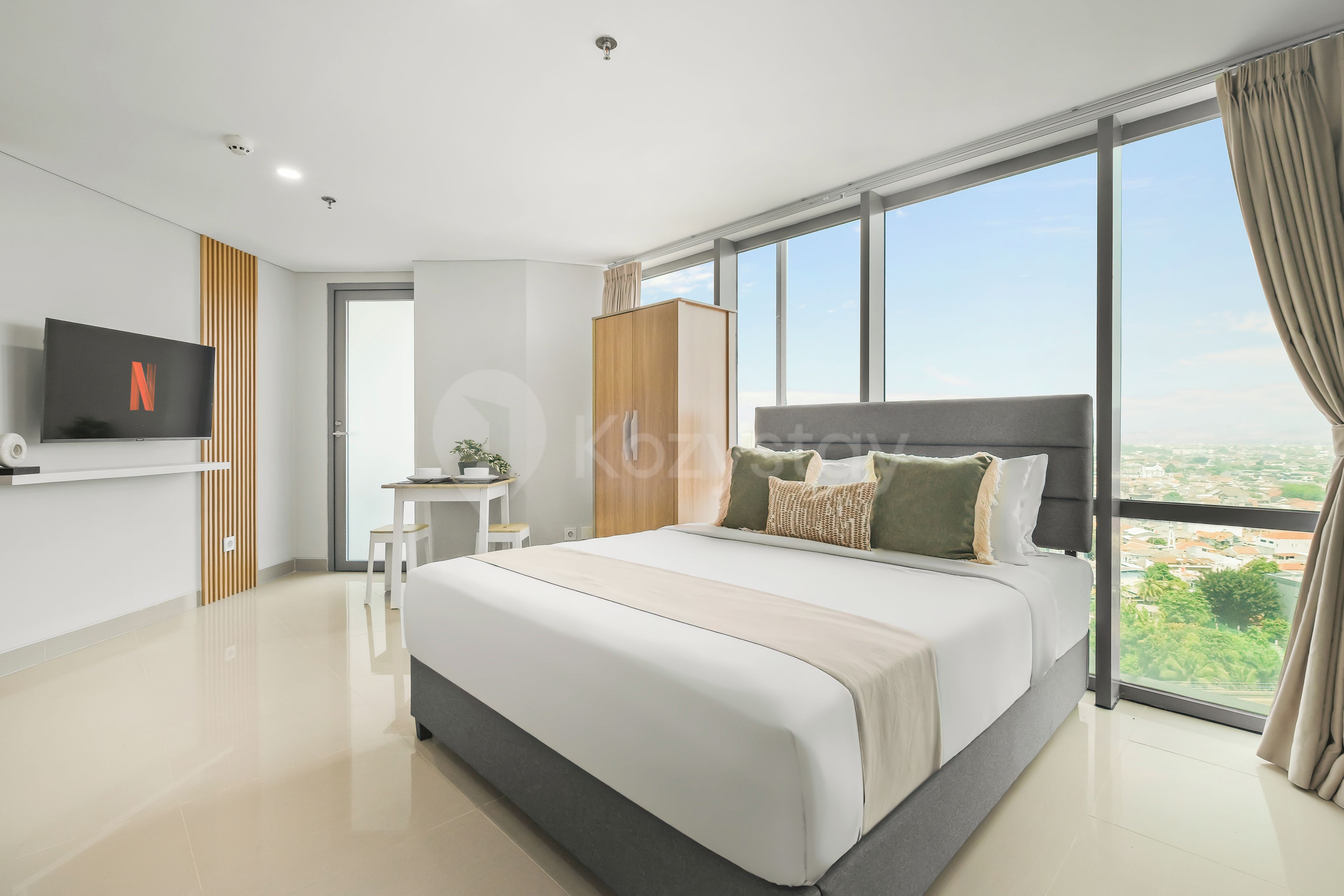 A modern bedroom with a large bed, a TV mounted on the wall, and a view of the city through large windows.