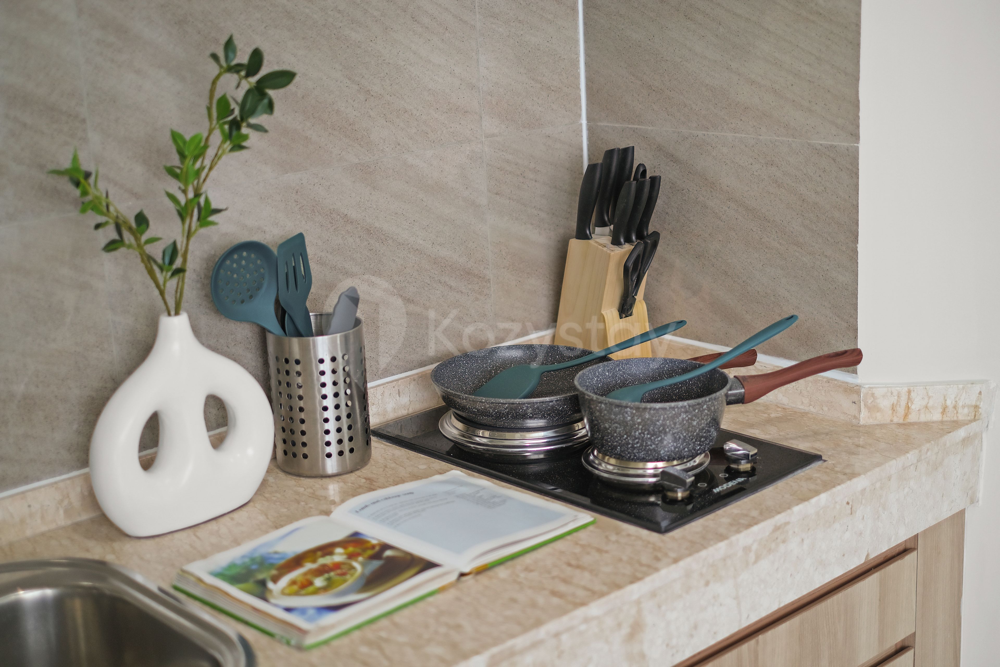 Una encimera de cocina con un jarrón blanco, un colador de metal, un juego de cuchillos de cocina y dos ollas sobre una estufa.