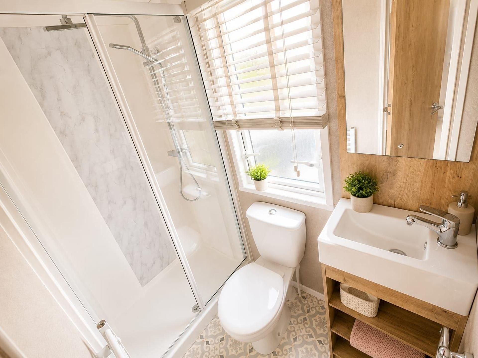 A modern bathroom with a glass shower enclosure, a toilet, a sink with a mirror, and a window with blinds.