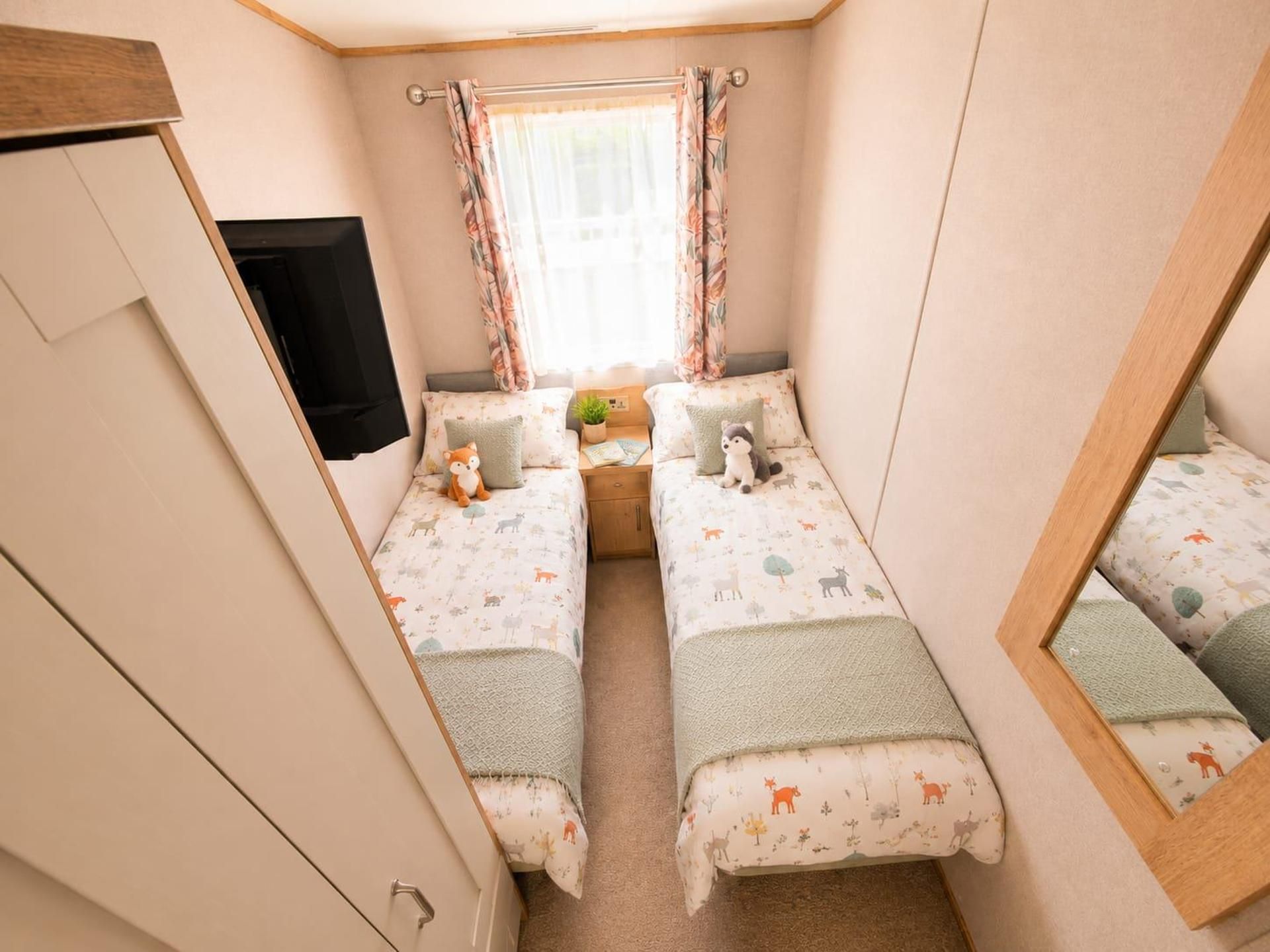 A compact bedroom with two single beds, a small bedside table, a TV, and a window with curtains.