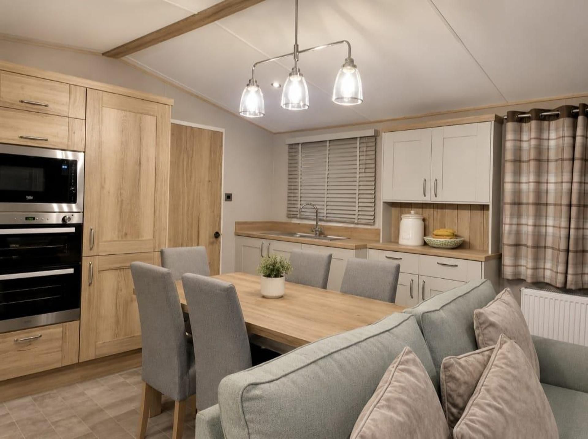 A modern kitchen with wooden cabinets, a built-in oven, a dining table with chairs, and a grey sofa.