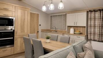A modern kitchen with wooden cabinets, a built-in oven, a dining table with chairs, and a grey sofa.