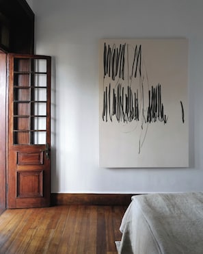A room with a wooden door, a large abstract painting on the wall, and a bed with a grey blanket. - Hakanto House (Antananarivo)