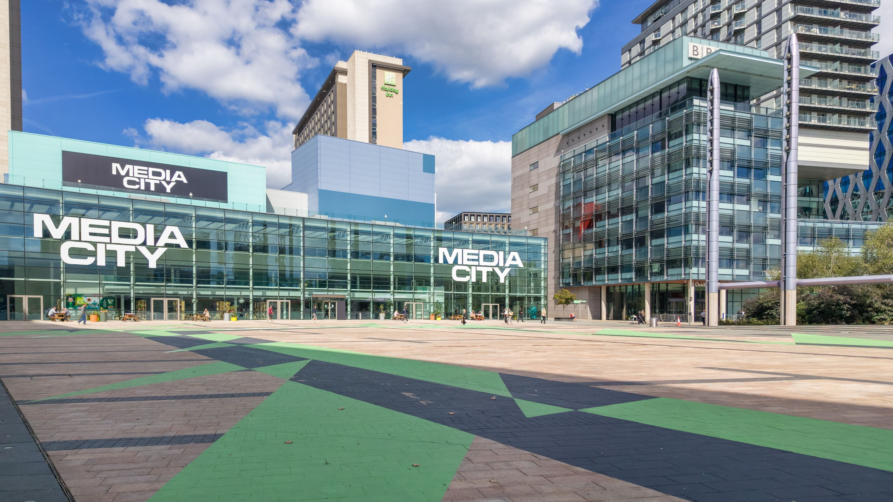 A modern building with 'MEDIA CITY' signage, featuring a glass facade and a green bike lane.