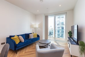 Un salon moderne avec un canapé bleu, un fauteuil gris, une petite table ronde et un téléviseur posé sur un meuble.