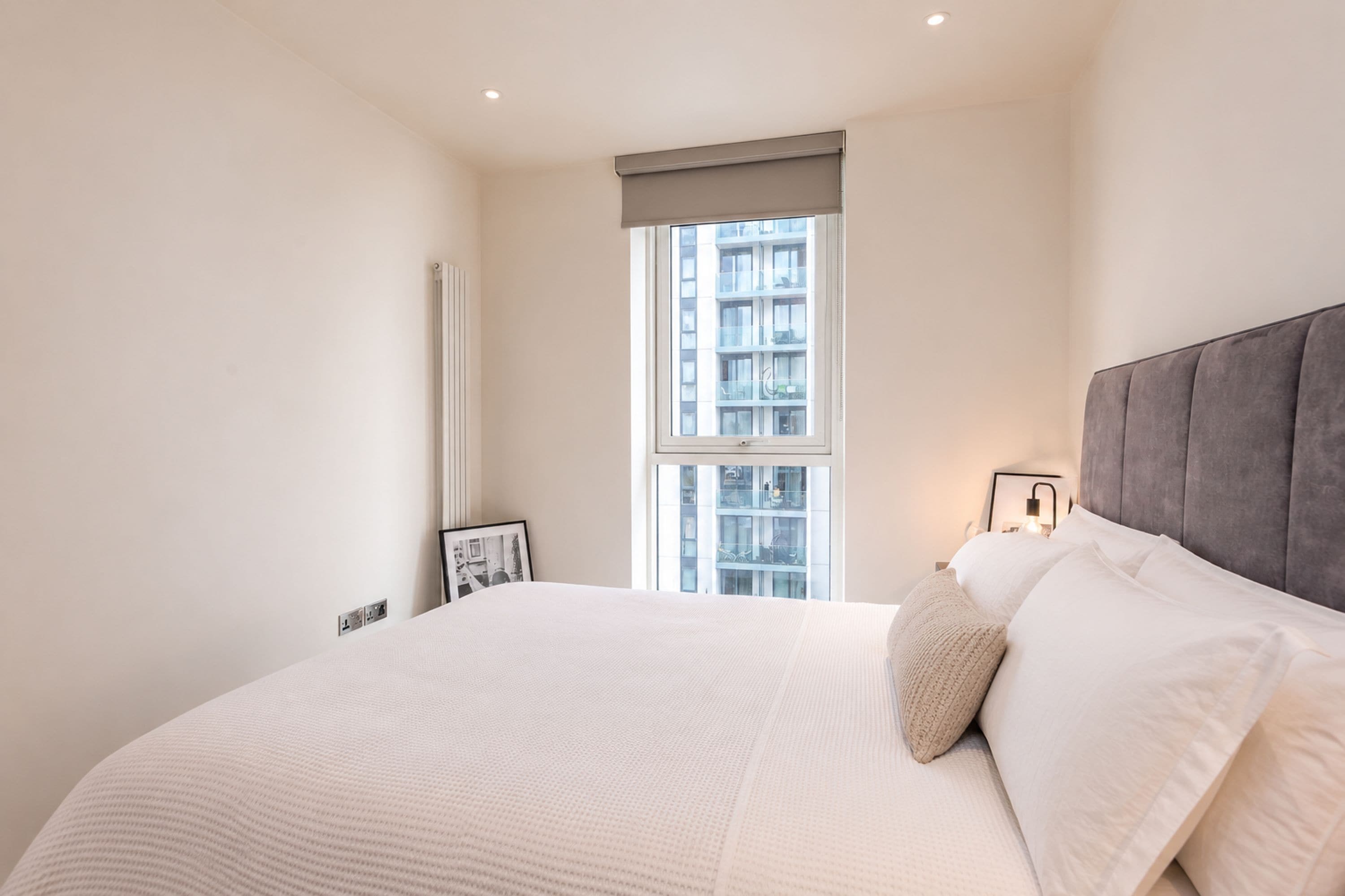 A bedroom with a large bed, a window with a view of buildings, a radiator, and a framed picture on the wall.