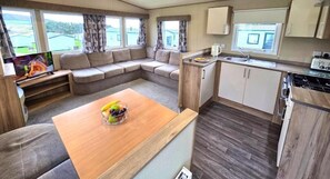 A compact RV interior with a living area, kitchenette, and a dining table with fruit. - Ruda Holiday Park (Croyde)
