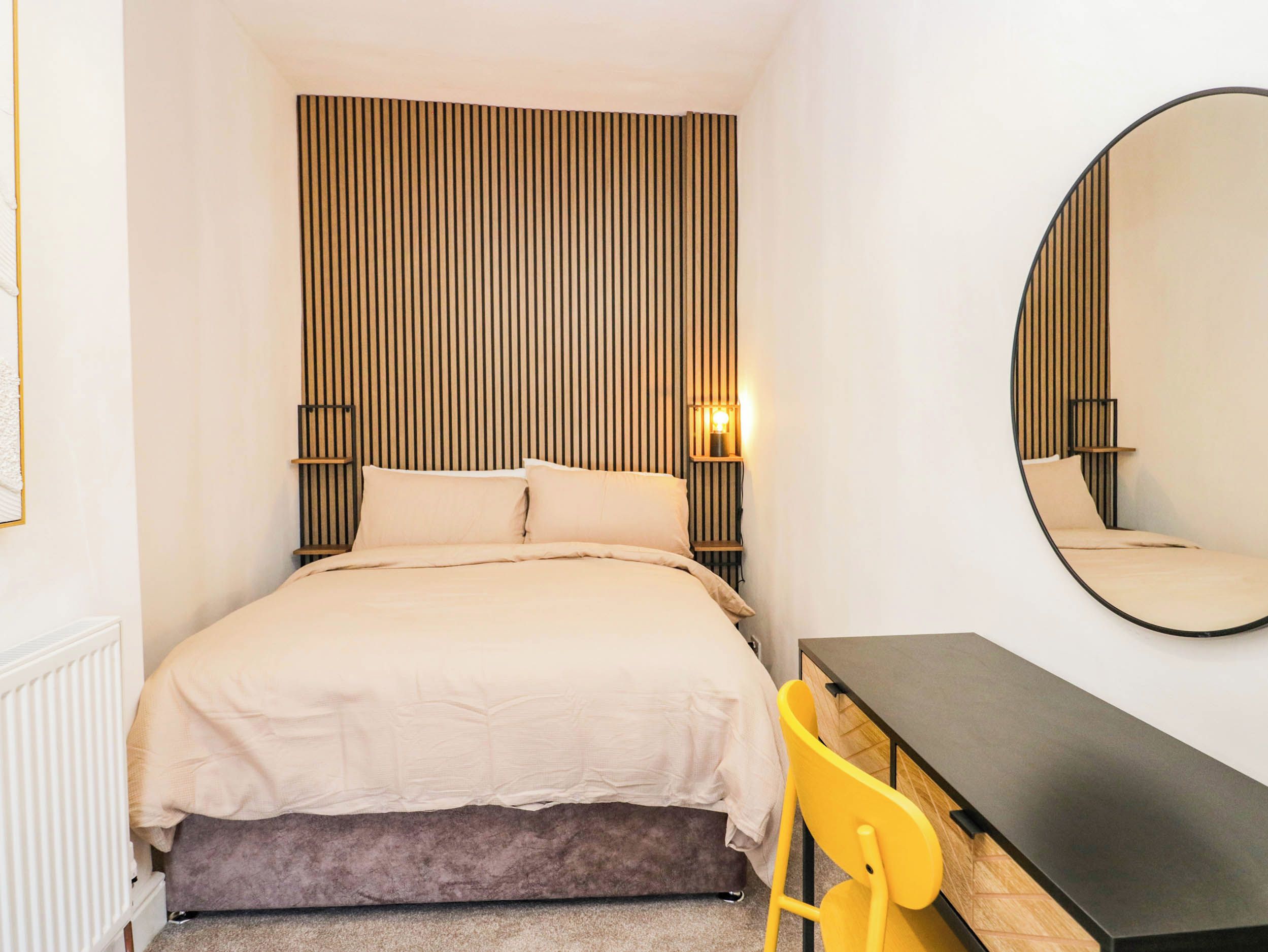 A bedroom with a bed, bedside table, chair, and a large mirror.