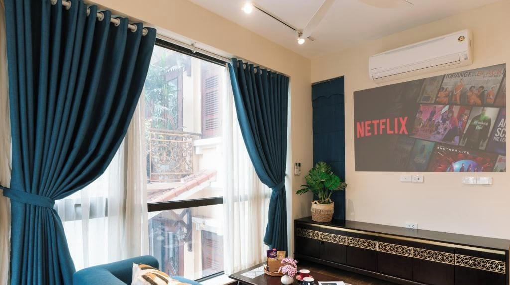 A room with a large window, a wall-mounted air conditioner, a flat-screen TV displaying a Netflix poster, and a small potted plant on a console table.