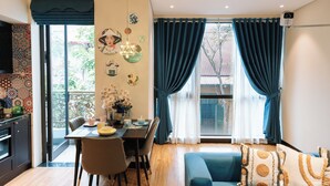 Ein modernes Wohnzimmer mit Essbereich, großen Fenstern mit Vorhängen und einer durch eine offene Tür einsehbarer Küche.