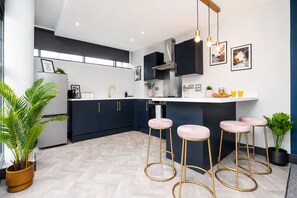 A modern kitchen with dark cabinetry, a white countertop, and three pink bar stools. - K Suites - 150m to New St Station w/ Balcony (Birmingham)