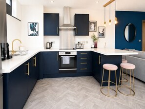 A modern kitchen with dark cabinetry, a white countertop, a stainless steel range hood, and a built-in oven. There are two bar stools with gold legs and a grey sofa in the adjacent living area. - K Suites - 150m to New St Station w/ Balcony (Birmingham)